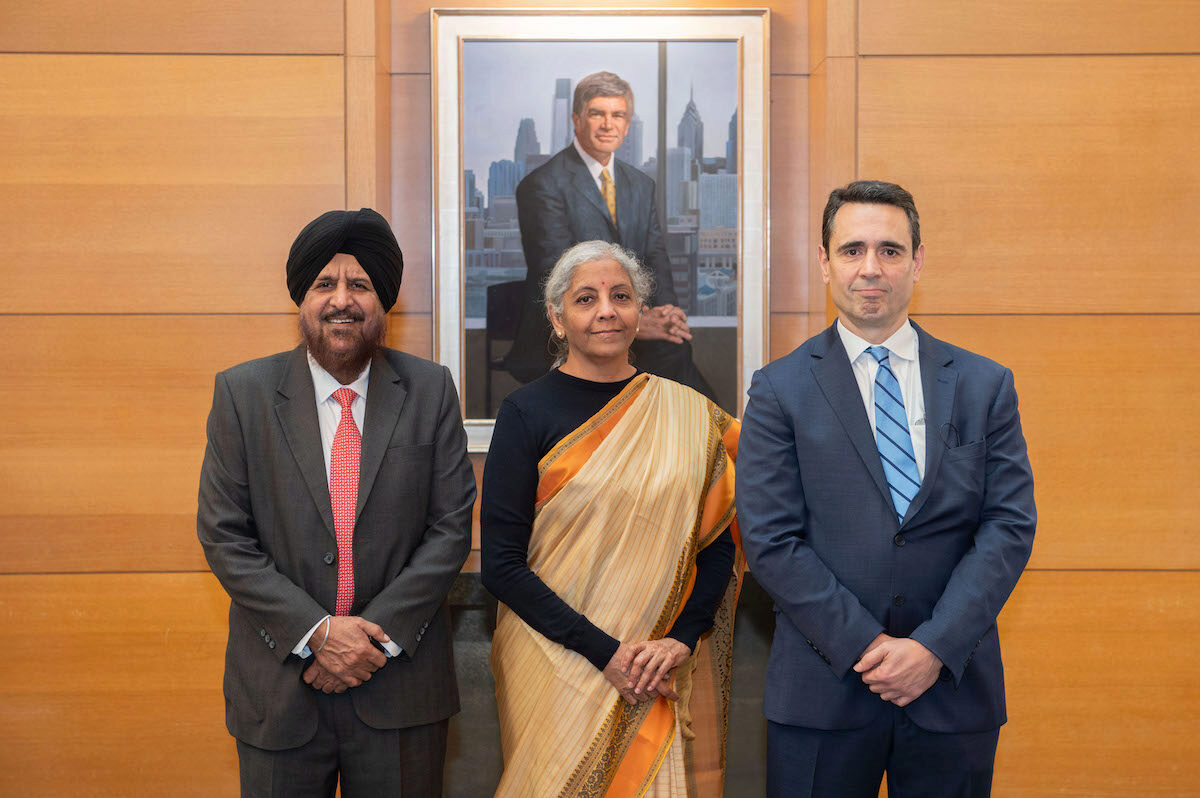 A small group of three people in formal attire stands in front of a wood-paneled wall and a framed portrait of a person in a suit.