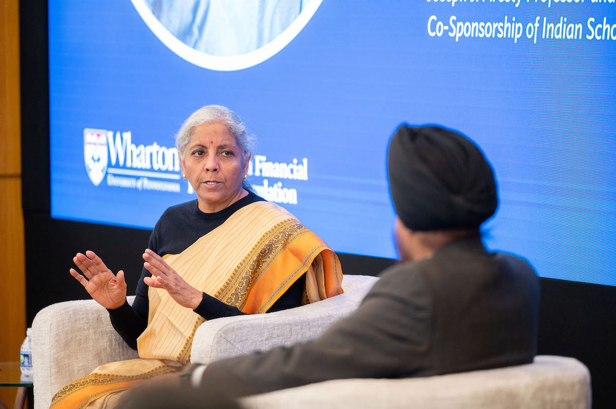 A person in a yellow saree is engaged in a discussion on stage with another individual. A large screen displaying text and the Wharton School logo is in the background.