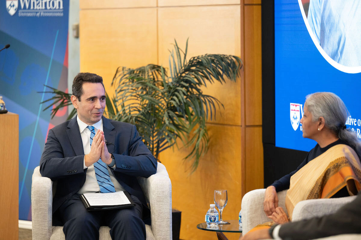 A formal discussion or interview setting with two people seated and engaged in conversation. One person gestures with hands together, wearing a suit, while the other, in traditional attire, listens attentively. A banner with "Wharton" is in the background,