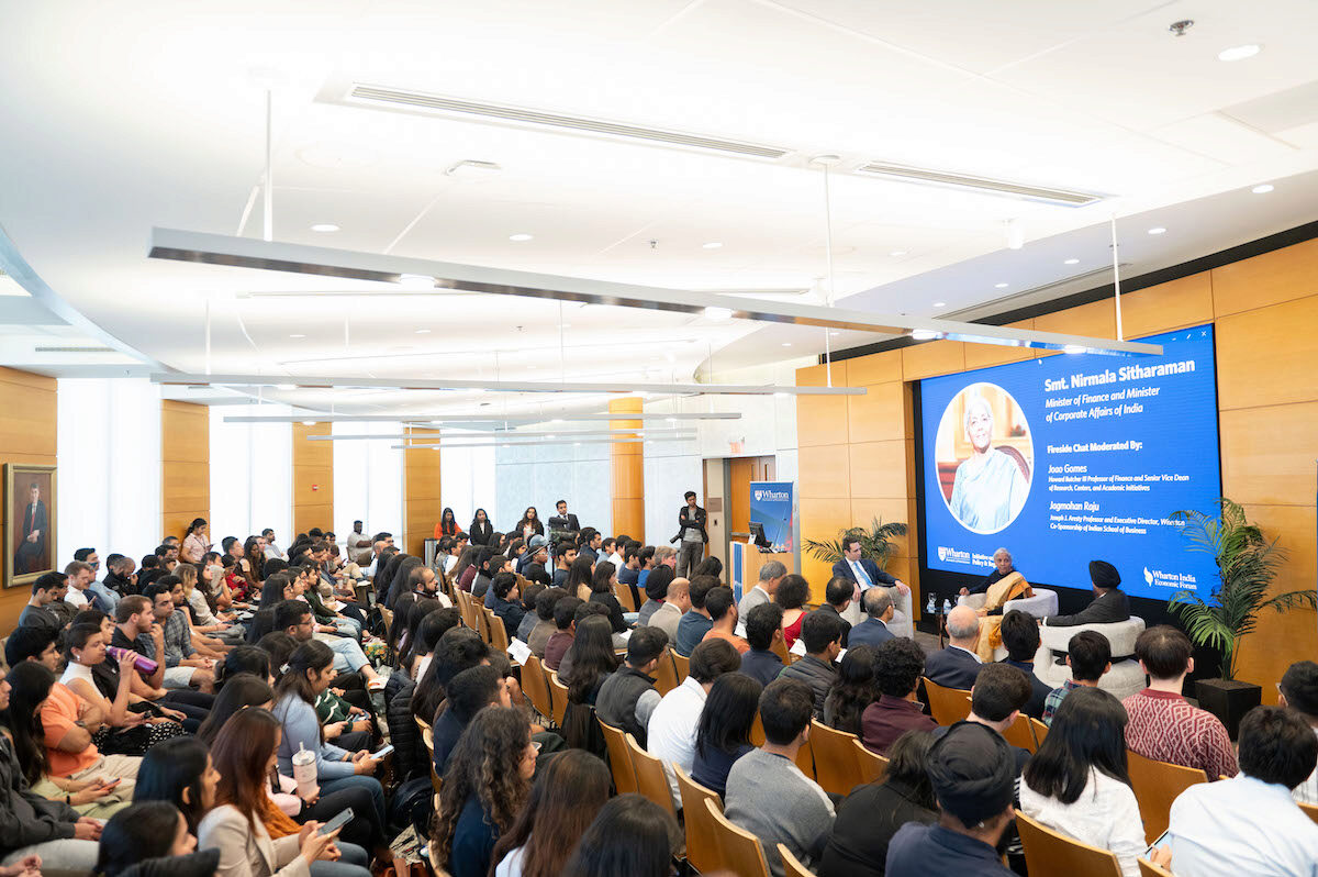 A large group of people is attending a fireside chat featuring a person sitting on stage beside a screen that reads "Smt. Nirmala Sitharaman, Minister of Finance and Minister of Corporate Affairs of India." The event appears to be at