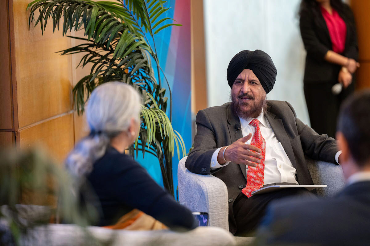 A formal discussion or interview setting with a person in a suit and turban gesturing while speaking to another person. A plant and a person with a microphone are in the background.