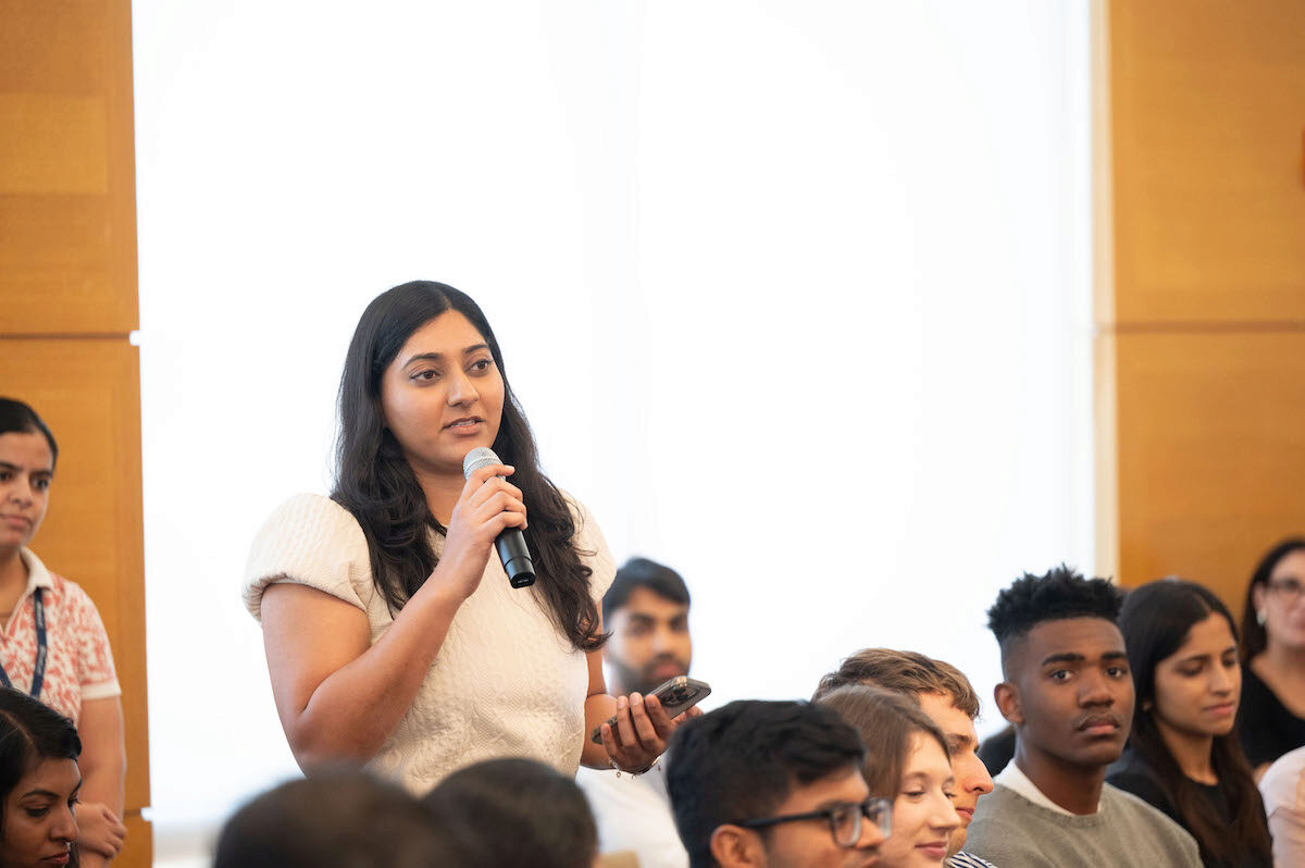 A person holding a microphone stands in a room filled with people seated, possibly at a conference or meeting. The atmosphere appears focused and attentive.