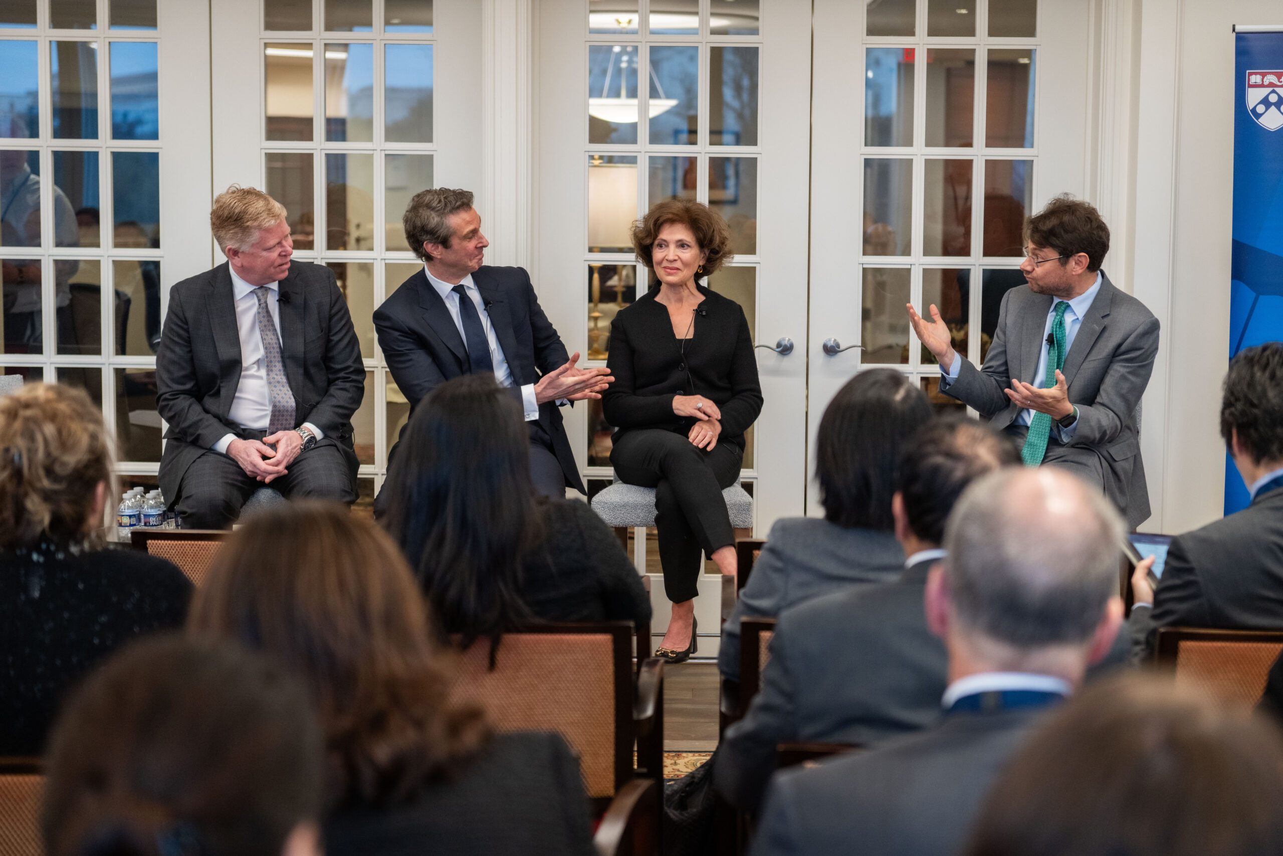 A panel discussion with four individuals seated in front of an audience. The group is engaged in conversation, dressed in formal attire, with attention focused on the speaker gesturing with hands. There is a banner beside the panelists.