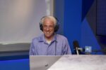 A podcast host smiles toward the viewer while wearing headphones