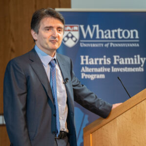 A person stands at a podium, in front of a sign that reads "Wharton Harris Family Alternative Investments Program"