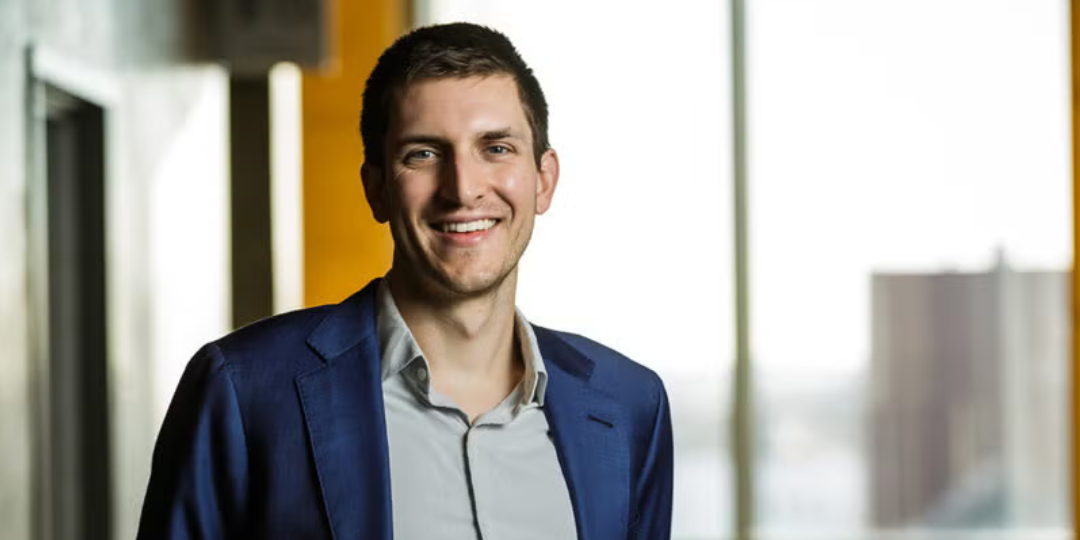 A person smiles in a sunlit room with large windows in the background