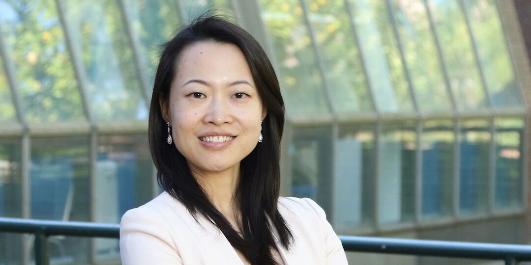 A person smiles toward the viewer in a sunlit room before a background of windows
